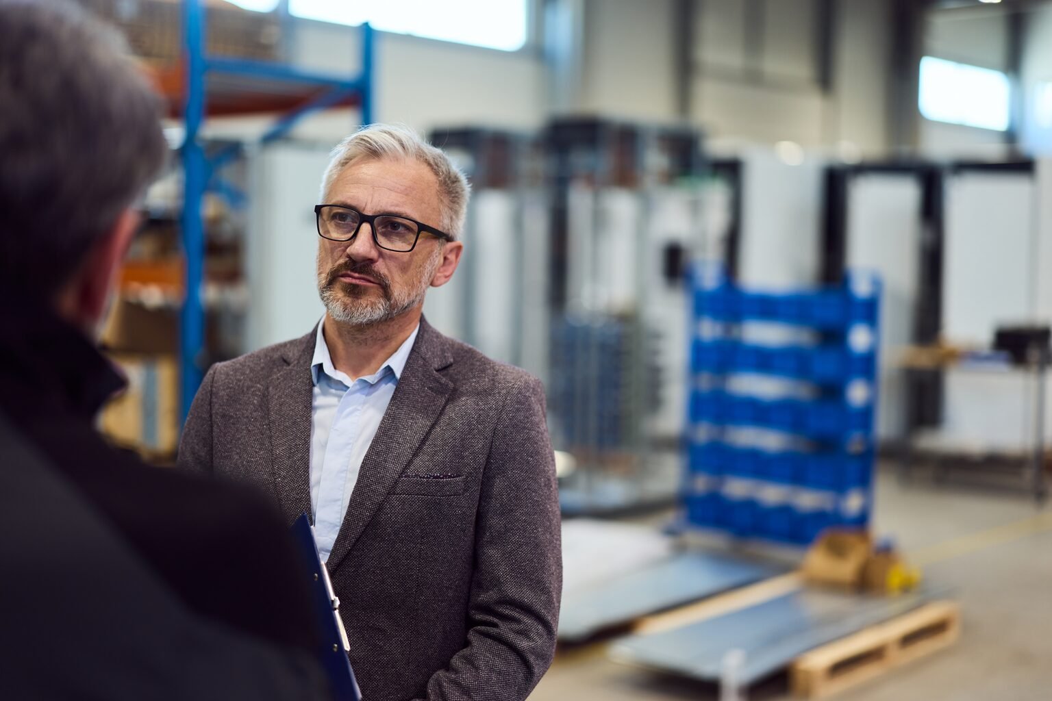 Inhaber eines mittelständischen Betriebs im Gespräch in der Produktionshalle, im Hintergrund Regale und Schaltschränke