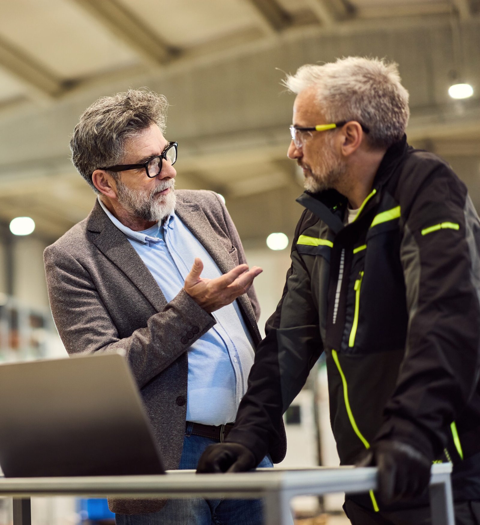 Zwei erfahrene Kollegen besprechen sich in der Fertigungshalle, einer in Arbeitsjacke, der andere im Sakko – ruhig und auf Augenhöhe.
