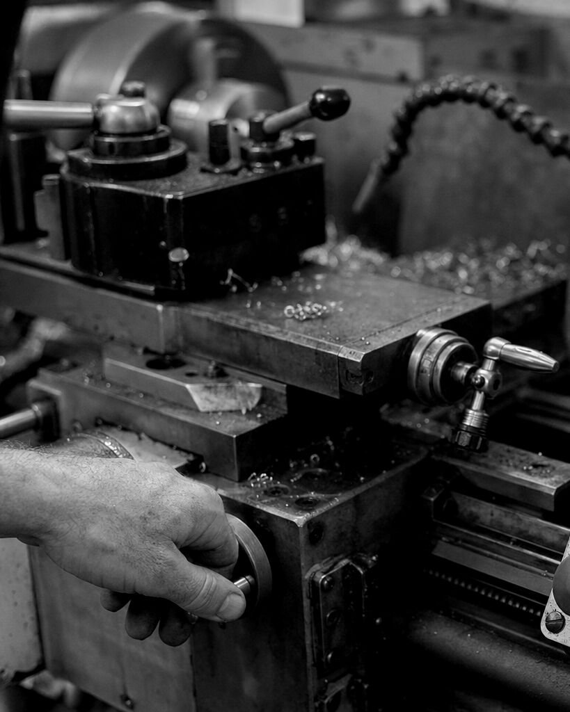 Schwarz-Weiß-Aufnahme einer Hand am Handrad einer Drehmaschine mit Spänen auf dem Schlitten