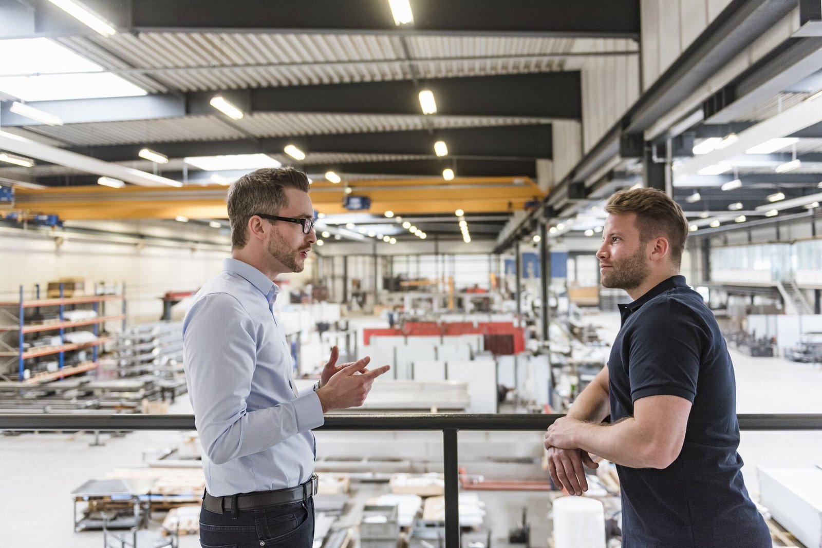 Zwei Männer im Gespräch an einem Geländer, unter ihnen eine helle Produktionshalle mit Regalen und Kranbahn