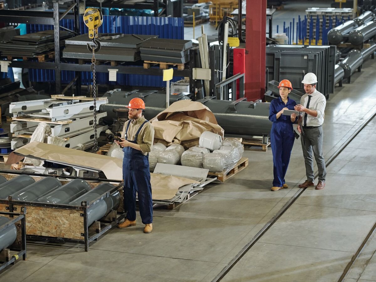 Blick von oben in eine Produktionshalle mit Stahlteilen, Paletten und drei Beschäftigten mit Schutzhelm